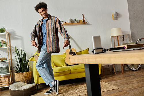 Young African American male blogger playing guitar, talking on phone camera in front of yellow couch.