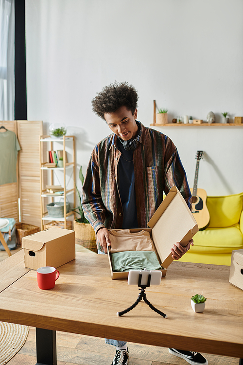 Young African American male blogger opening a box in living room while talking on phone camera.