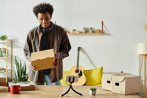 A young African American male blogger opens a box while talking on a phone camera in his living room.