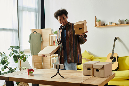 Young African American man holding cardboard box while vlogging in living room.