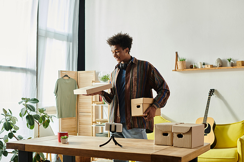 A young African American male blogger is holding a box in front of a table while talking on a phone camera.