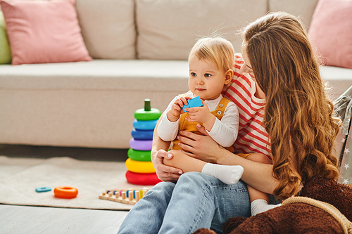 A young mother lovingly holds her toddler daughter while sitting on a cozy couch at home.