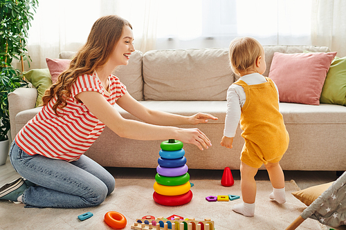 A young mother and her toddler daughter laughing and playing in a cozy living room.