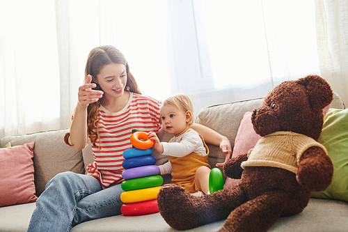 A young mother sits on a couch with her baby daughter and a teddy bear, sharing a tender moment of love and togetherness.