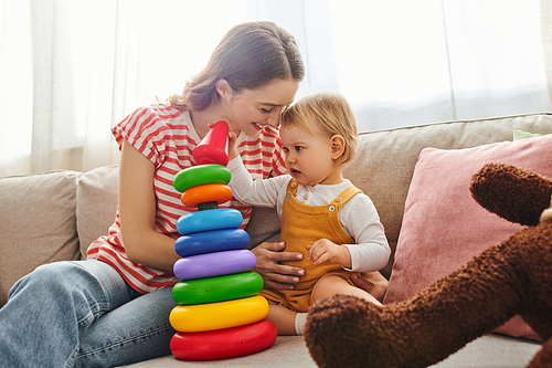 A young mother and her toddler daughter laughing and playing together on a couch in a cozy home setting.