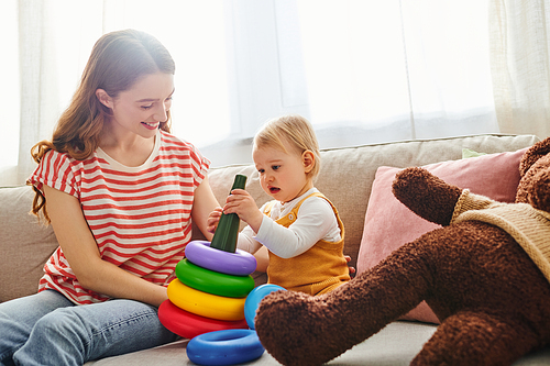 A young mother sits on a cozy couch, tenderly playing with her smiling toddler daughter in a heartwarming moment of love.