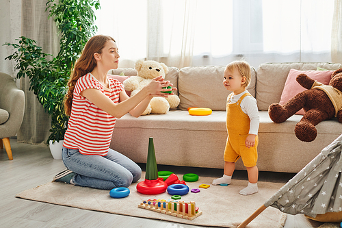 A young mother engages playfully with her daughter amidst toys in a cozy living room.