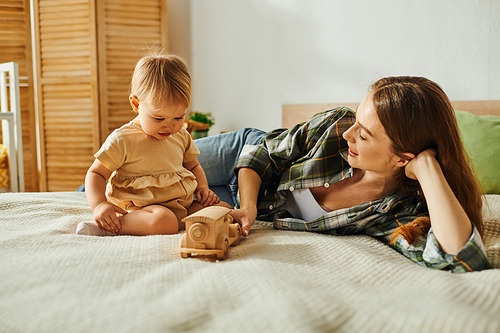 A young mother lays on a bed with her baby daughter, both smiling, while a toy rests beside them.