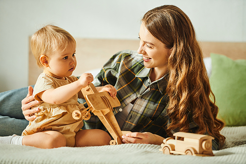 A young mother lovingly interacts with her toddler daughter while playing on a cozy bed at home.