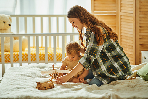 A young mother joyfully plays with her toddler daughter on a cozy bed, sharing smiles and laughter in a heartwarming scene.