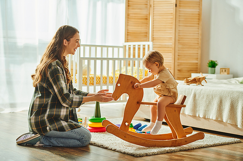 A young mother happily plays with her toddler daughter on a wooden rocking horse, creating sweet memories at home.