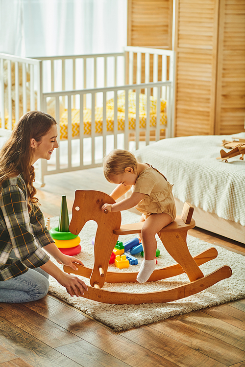 A young mother joyfully plays with her toddler daughter on a traditional wooden rocking horse, bonding and creating cherished memories.