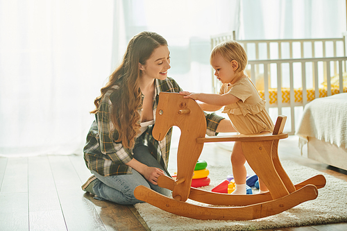 A young mother joyfully plays with her toddler daughter on a wooden rocking horse in their cozy home.