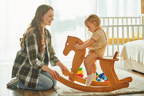 A young mother happily plays with her toddler daughter on a rocking horse at home, bonding and creating special memories together.