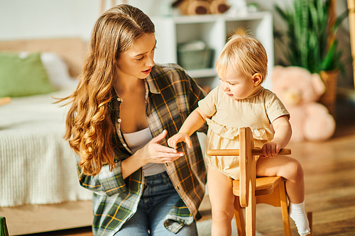 A young mother sitting in a chair beside her toddler daughter, sharing a warm moment together at home.