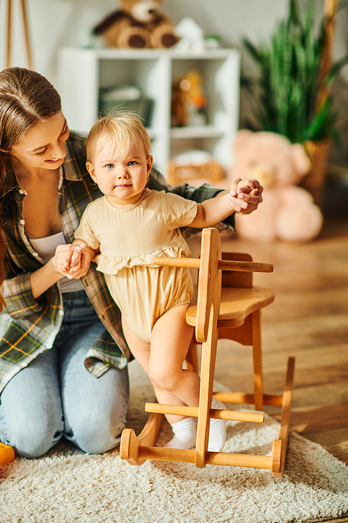 A young mother kneels next to her toddler daughter sitting in a high chair, sharing a precious moment together at home.