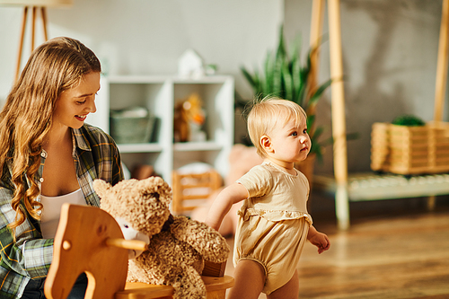 A young mother sits in a rocking chair holding a teddy bear next to her baby daughter, sharing a sweet moment together.