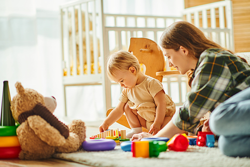 A young mother and her toddler daughter cheerfully engage with toys on the floor, building a strong, loving connection.