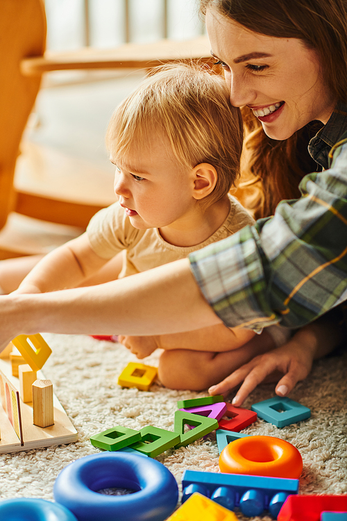 A young mother and her toddler daughter are happily playing with toys on the floor at home.