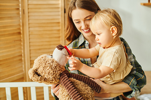 A young mother tenderly holds her baby daughter in one arm while a teddy bear sits in the other, creating a scene of love and comfort.