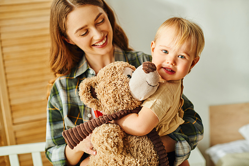 A young mother holding her baby daughter in her arms while the toddler cuddles a teddy bear, showing love and bond.