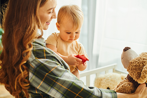 A young mother lovingly holds her baby daughter and a teddy bear, creating a heartwarming scene of affection and bonding.