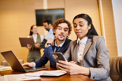 Man and woman of different ethnicities sitting at a table, engrossed in a cell phone screen.