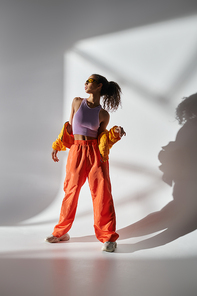 A young woman in a studio wears a yellow jacket, purple tank top, orange pants, and sunglasses, exuding coolness.