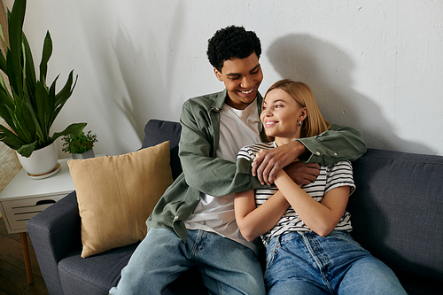 A young, multicultural couple embraces on a couch in a modern apartment.