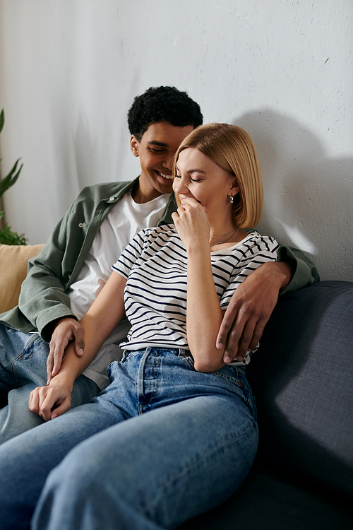 A young interracial couple sits on a sofa, sharing a laugh and a moment of intimacy in their modern apartment.