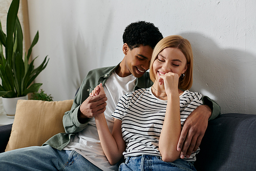 A young interracial couple sits on a couch in a modern apartment, laughing together.