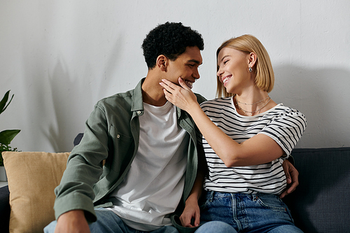 A young, multicultural couple sits on a couch in a modern apartment, sharing a tender moment of intimacy and affection.