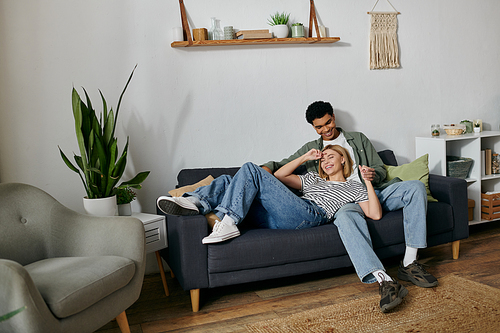 A young multicultural couple laughs and enjoys each others company on a comfortable couch in their modern apartment.