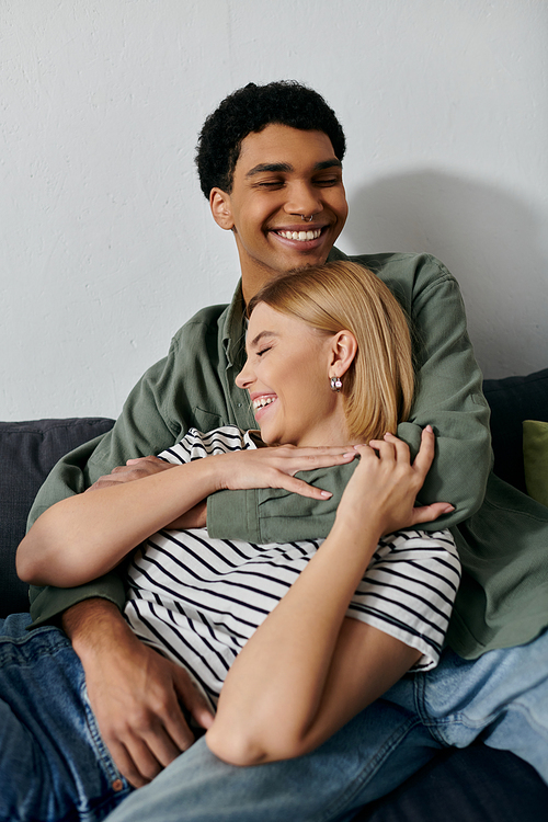 A young couple, one man and one woman, laugh and embrace on a couch in a modern apartment.