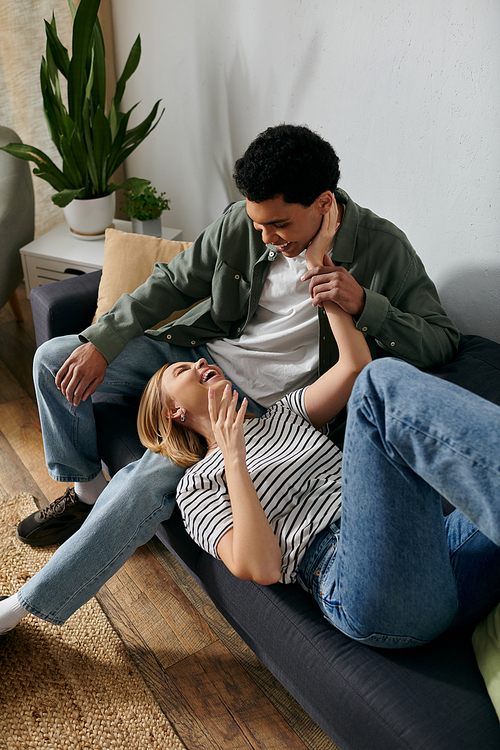 A young, multicultural couple shares a moment of laughter and affection while relaxing on a couch in their modern apartment.