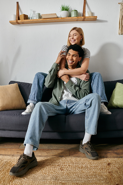 A young interracial couple laughs together on a couch in a modern apartment.