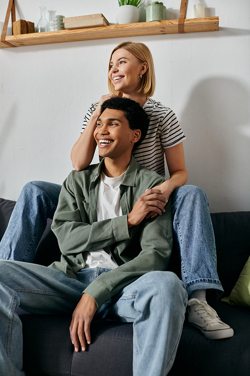 A young multicultural couple sits on a couch in a modern apartment, smiling and enjoying each others company.