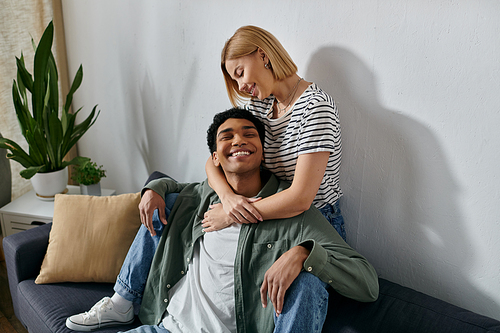 A young interracial couple relaxes on a couch in a modern apartment, enjoying a moment of intimacy and affection.