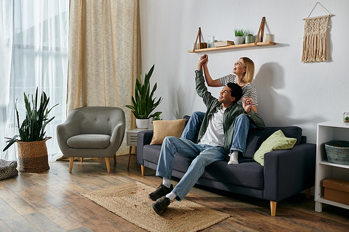 A young multicultural couple celebrates with raised arms, sitting on a sofa in a modern apartment.