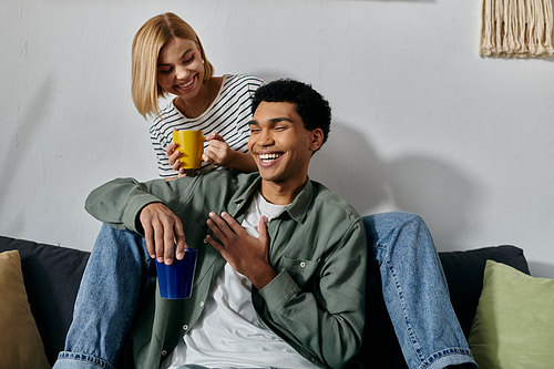 A young interracial couple relaxes on a couch in a modern apartment, enjoying a cup of coffee and each others company.