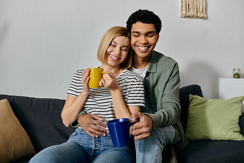 A young, interracial couple sits on a couch in their modern apartment, sharing a moment of cozy intimacy and laughter.