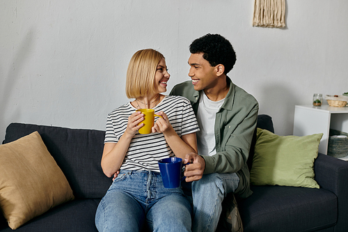 A young multicultural couple sits on a couch in a modern apartment, enjoying coffee together.