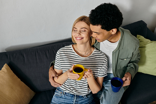 A young interracial couple enjoys coffee together on a comfortable couch in a modern apartment.