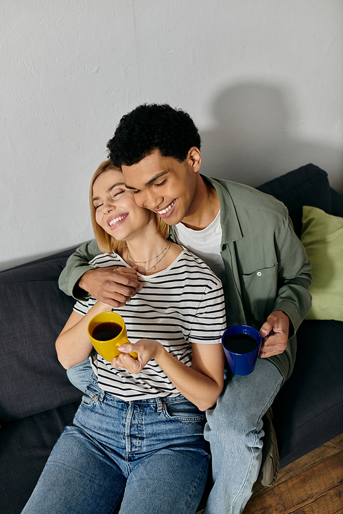 A young interracial couple relaxes on a couch in a modern apartment, sharing coffee and a cozy moment together.