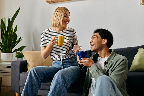 A young multicultural couple sits on a couch in their modern apartment, enjoying coffee and conversation.