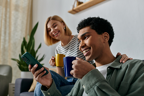 A young multicultural couple enjoys a relaxing moment in their modern apartment, laughing and sharing a cup of coffee.