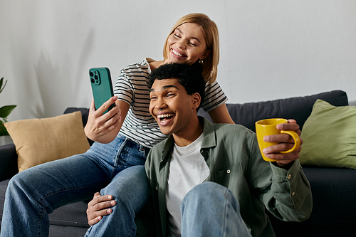 A young multicultural couple smiles and laughs as they take a selfie with a phone in their modern apartment.