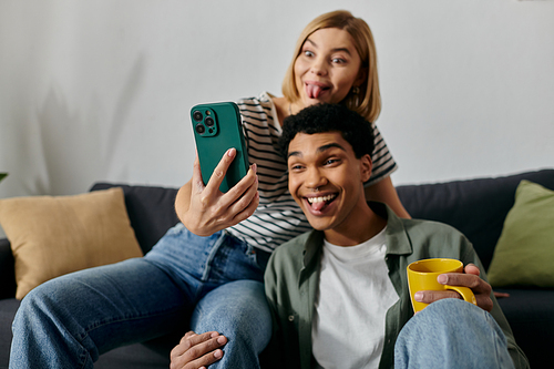 A young multicultural couple sits on a couch in their apartment, taking a silly selfie together.