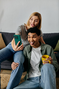 A young multicultural couple takes a selfie together while making funny faces in their modern apartment.