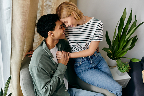 A young multicultural couple sits together on a couch in their modern apartment, sharing a tender moment.